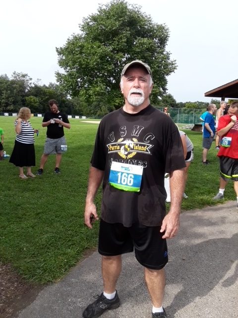 A man in a black USMC t-shirt and running shorts stands with a race bib numbered 166 at a community run event