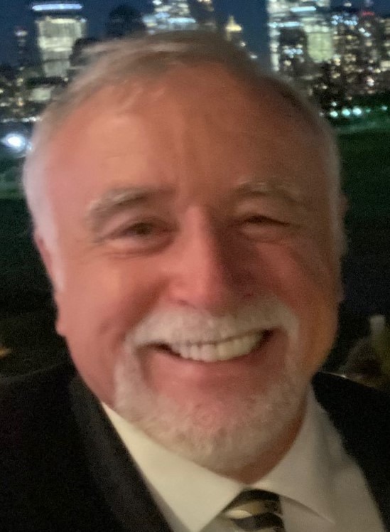 Smiling man with gray hair and a white beard, wearing a suit, against a nighttime city skyline