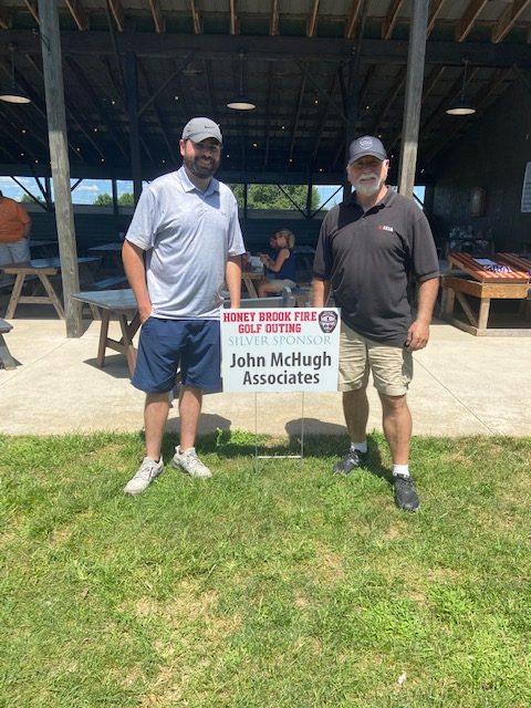 Two men stand next to a sign promoting the Honey Brook Fire Golf Outing, sponsored by John McHugh Associates