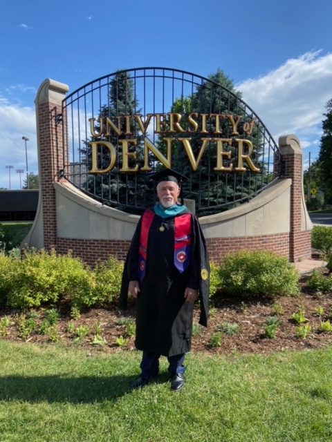 Graduating student in cap and gown stands proudly in front of the University of Denver sign